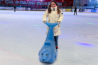 Kind fährt mit blauer Eislauf-Laufhilfe auf einer Eislaufbahn in der Halle.